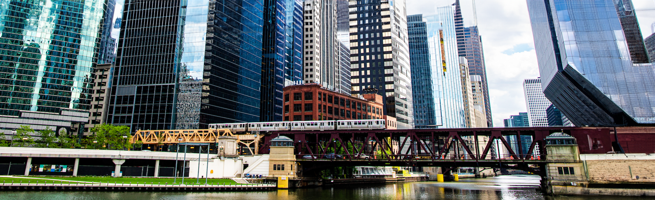 A scene of downtown Chicago, including the Chicago River, the Riverwalk, the elevated CTA train on a bridge over the river, and skyscrapers.